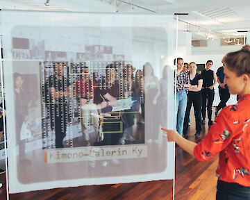 Lilly Lulay steht vor Besuchenden in ihrer Ausstellung. Sie zeigt von hinten auf einen bedruckten Stoff.  © buerofuerkunstdokumentation