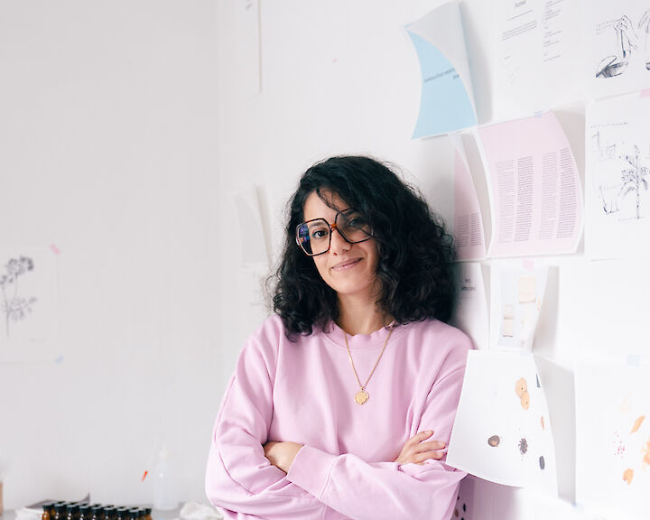 Portrait of Btihal Remli. She is wearing glasses and a pink sweater and is leaning against the wall of her studio.