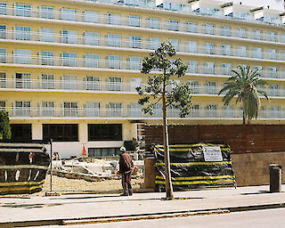 [Eine Person bleibt vor einem sehr großen Hotel in dem Urlaubsort Lloret de Mar, Mallorca, stehen. Die Hotelfassade füllt das gesamte Bild aus und zeigt sechs Stockwerke. Sichtbar sind zwanzig aneinander gereihte Fenster und Balkone. Das Hotel ist geschlossen. Ein Bauzaun grenzt das Grundstück ab. An dem Zaun hängt ein Schild mit der Telefonnummer einer Firma.] Lloret de Mar ©Caroline Brünen