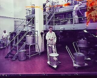 Arbeiter in einer Papierfabrik, die Maschinen bedienen und mit großen Fässern hantieren. Die Szene zeigt Metallgeräte und Treppen auf einem gefliesten Boden, der von einem violetten Farbton überlagert wird. Fotograf:in unbekannt, Barytküche, 17.12.1956, Farbfotografie, Museum Ludwig, Agfa Werbearchiv © Fotograf*in unbekannt, Agfa Werbearchiv, Museum Ludwig