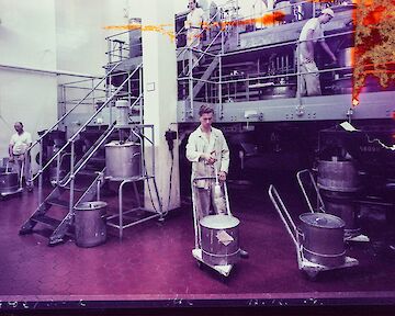 Arbeiter in einer Papierfabrik, die Maschinen bedienen und mit großen Fässern hantieren. Die Szene zeigt Metallgeräte und Treppen auf einem gefliesten Boden, der von einem violetten Farbton überlagert wird. Fotograf:in unbekannt, Barytküche, 17.12.1956, Farbfotografie, Museum Ludwig, Agfa Werbearchiv © Fotograf*in unbekannt, Agfa Werbearchiv, Museum Ludwig