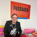 Sabine Voggenreiter sitzt an einem dunkeln Tisch. Sie trägt einen Blazer mit silberner Brosche und hat die braunen Haare zusammengebunden. Hinter ihr an der Wand hängt ein großes Poster der PASSAGEN von 1990.