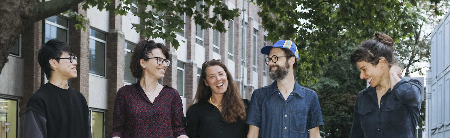 The 5 Artist Meets Artist artists stand smiling on a cobbled street under green trees. Elena Efeoglou has a dachshund on a leash.