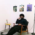 The artist is sitting on a low chair in front of a white wall. He is wearing dark clothing and has one arm propped up. Behind him hang three printed photos.