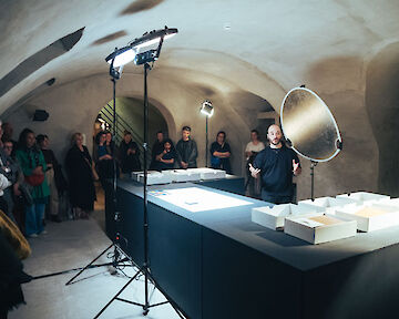 Pablo Lerma stands behind a table in front of visitors and speaks into a microphone. In front of him are empty boxes.  © buerofuerkunstdokumentation