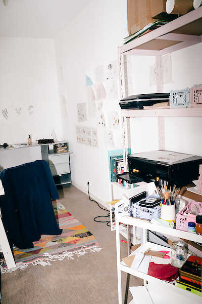 Blick in ein Atelier. Man sieht ein Regal mit verschiedenem Werkzeug und an der Wand hängen viele Bilder.
