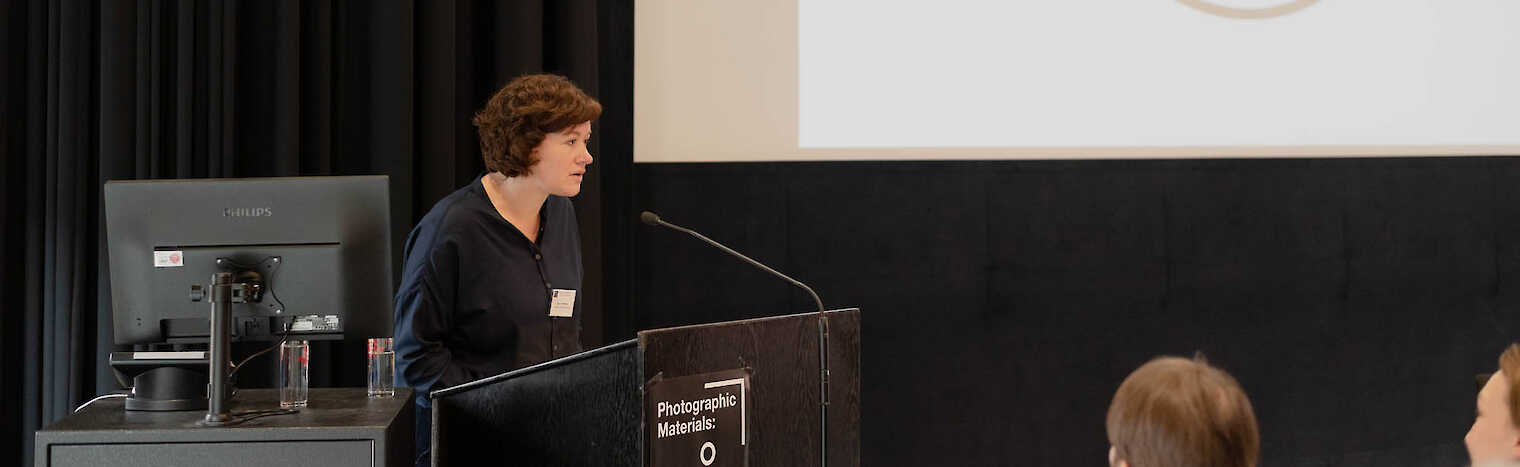 Inga Schneider stands at a lectern. Behind her on a screen you can see the Artist Meets Archive logo.