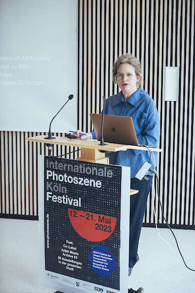 A woman stands at a lectern. The poster for the Photoszene Festival 2023 hangs in front of her.