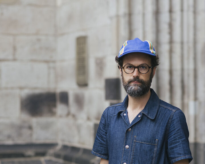 Andres Galeano in einem Jeans-Overall, mit einer Cappy und Brille. eine Hand in der Hosentasche.  ©buerofuerkunstdokumentation