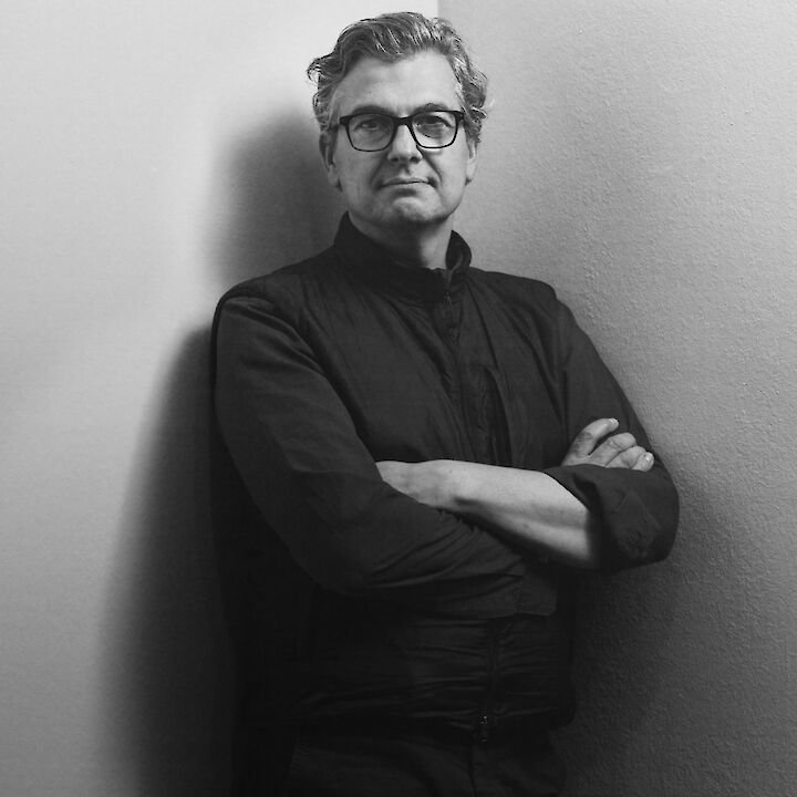 Portrait of Felix Hoffmann in black and white. He is leaning against a wall with his arms folded and looking into the camera. He is wearing glasses and a dark shirt. Felix Hoffmann © Michael Dooney