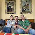 Three people sit on a couch smiling, surrounded by two paintings with ornate frames on a wall. A single flower in a vase sits on a colorful tablecloth in the foreground. The scene is set in a cozy living room.