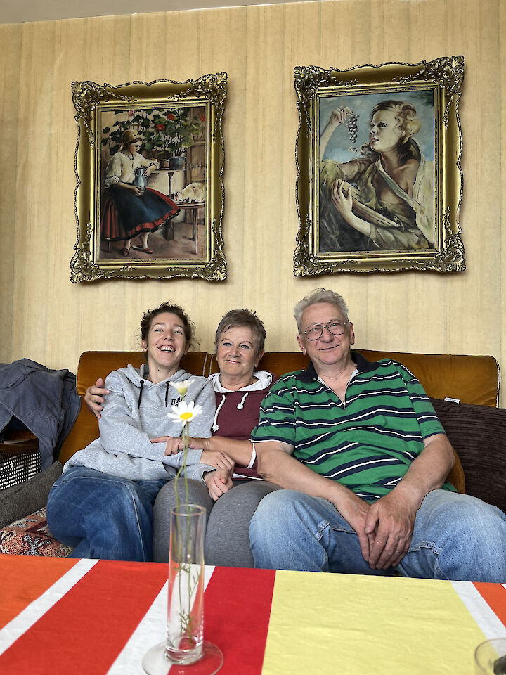 Drei Personen sitzen lächelnd auf einer Couch, umgeben von zwei Gemälden mit verzierten Rahmen an einer Wand. Eine einzelne Blume in einer Vase steht auf einem bunten Tischtuch im Vordergrund.