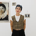 The artist stands in her exhibition. She is wearing red lipstick and a vest.