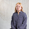The artist, a person with long blond hair and glasses is sitting against a backdrop of woven fabric art.