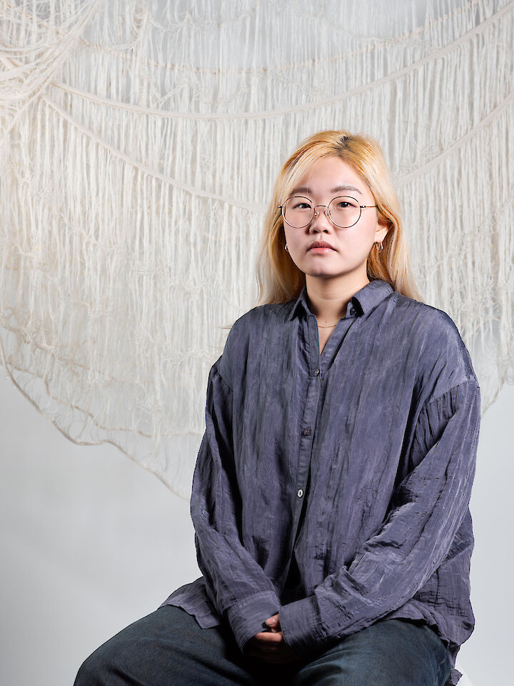 Die Künstlerin, eine Person mit langem blondem Haar und Brille, sitzt vor einem Hintergrund aus gewebten Stoffkunstwerken.  © Vivian Hötter