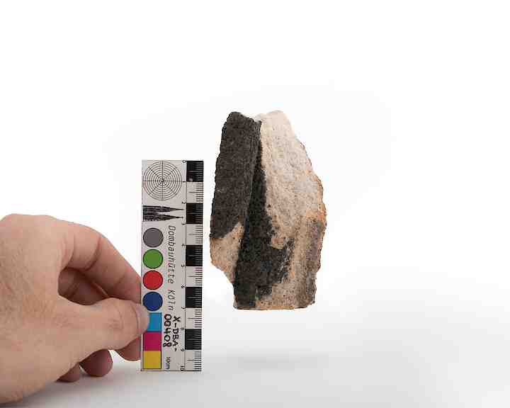 A hand holds a color calibration card next to a rough stone with black and light brown sections, demonstrating its size and texture. Andrés Galeano, X-DBA-00408 #1, 2024 © Andrés Galeano