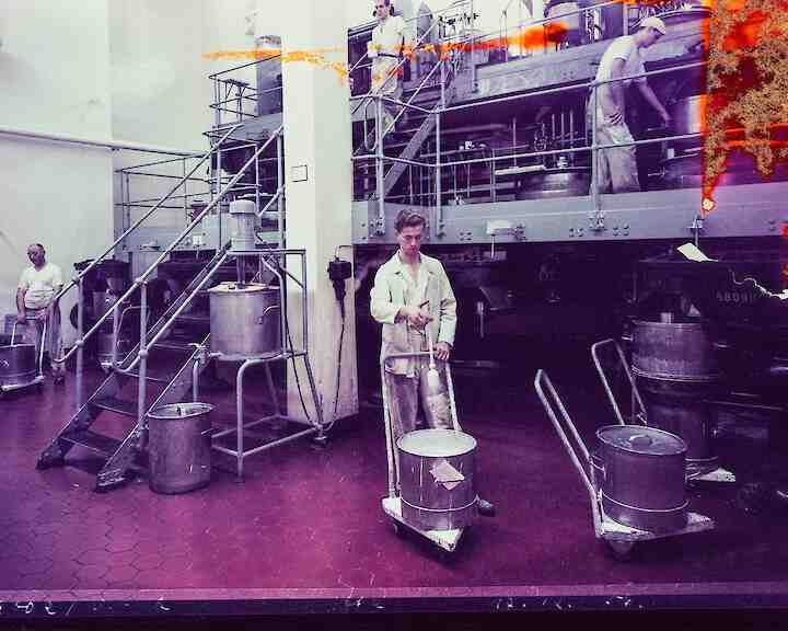 Workers in a paper factory operating machinery and handling large barrels. The scene includes metal equipment and staircases against a tiled floor, with a purple hue overlay. Fotograf:in unbekannt, Barytküche, 17.12.1956, Farbfotografie, Museum Ludwig, Agfa Werbearchiv © Fotograf*in unbekannt, Agfa Werbearchiv, Museum Ludwig