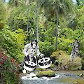 View of a river in the jungle. A monkey in black and white wears human clothes and two wreaths of beer and walks towards the river. Another comes through the bushes on a motorcycle. Both chimpanzees appear to have been collaged from other photos.