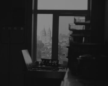 Schwarz-Weiße Innenaufnahme aus einem dunklen Raum, mit Blick auf die Skyline einer Stadt durch ein Fenster. The View of St. Michaels Cathedral, Qingdao, 2024 © Jimmi Wing Ka Ho