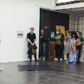 Eine Person in blauer Hose steht vor einer Gruppe von Menschen in einem Ausstellungsraum. Sie erzählt ihnen etwas.