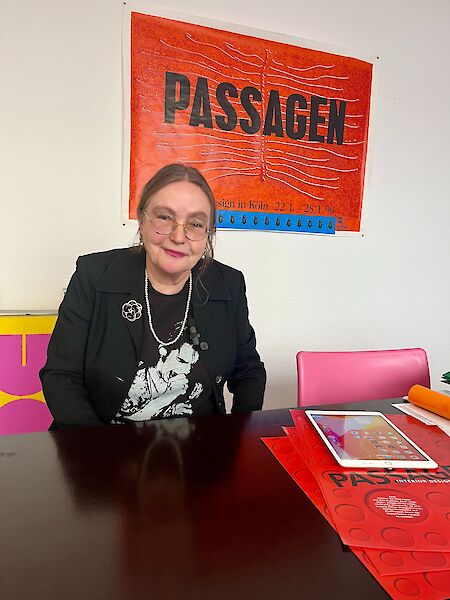 Sabine Voggenreiter sits at a dark table. She wears a blazer with a silver brooch and has tied her brown hair back. Behind her on the wall hangs a large poster of PASSAGEN from 1990.