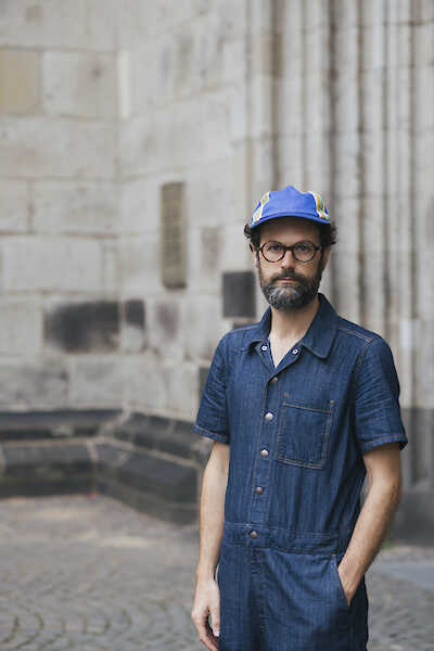 Andres Galeano in einem Jeans-Overall, mit einer Cappy und Brille. eine Hand in der Hosentasche.