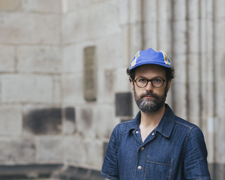 Andres Galeano in einem Jeans-Overall, mit einer Cappy und Brille. eine Hand in der Hosentasche.  © buerofuerkunstdokumentation